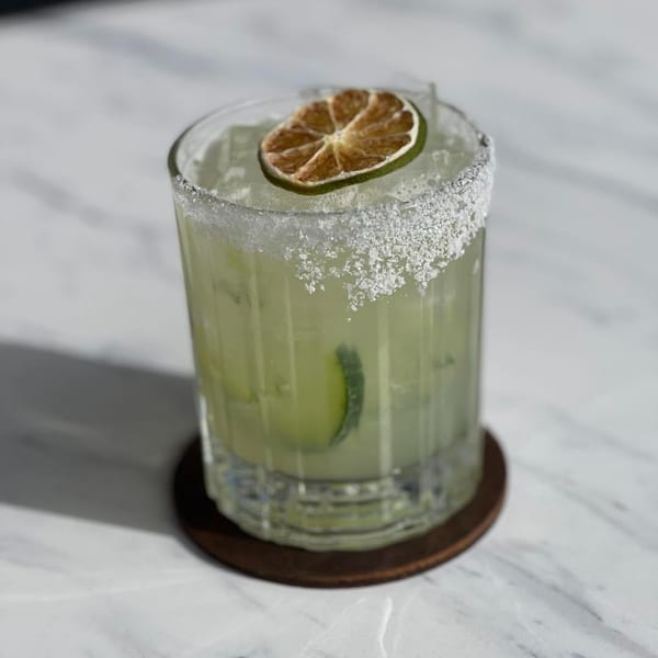 A light green cocktail with a salted rim, garnished with a dried lime slice and cucumber.
