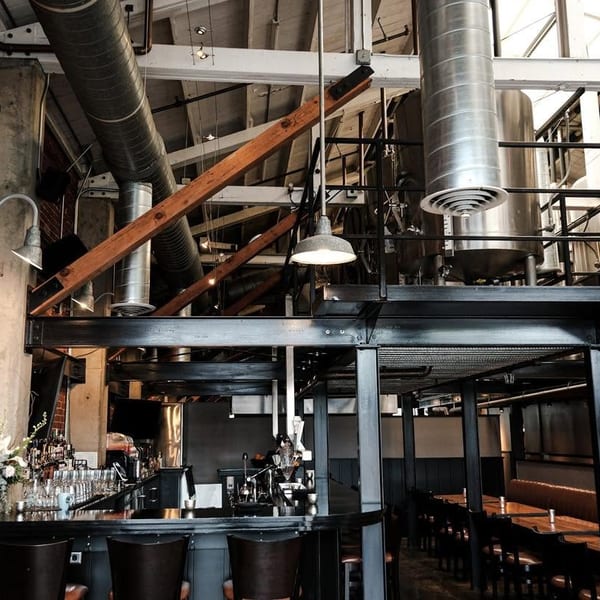 Industrial-chic restaurant interior with exposed brick, a bar, dining area, and brewing tanks overhead.