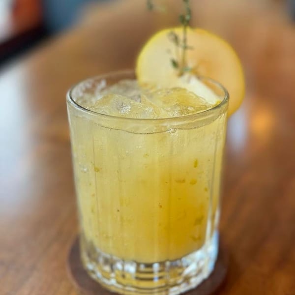 A vibrant yellow cocktail with ice, garnished with a melon slice and herbs on a wooden table.