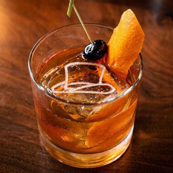 Old Fashioned cocktail with a top hat ice cube, orange peel, and cherry on a wooden table.