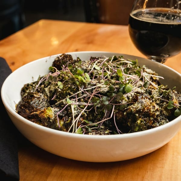 A white bowl of roasted Brussels sprouts topped with microgreens and parmesan, next to a dark beer.