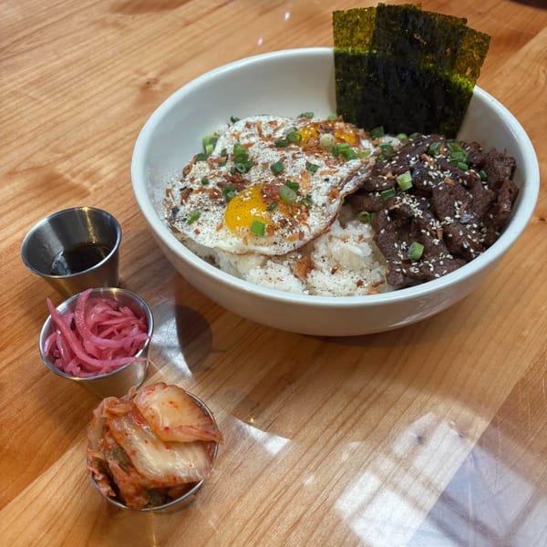 Korean BBQ beef bowl with fried eggs, rice, seaweed, pickled onions, and kimchi on a wooden table.