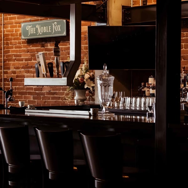 A dark, inviting bar with a brick wall, "The Noble Fox" sign, beer taps, and liquor bottles.
