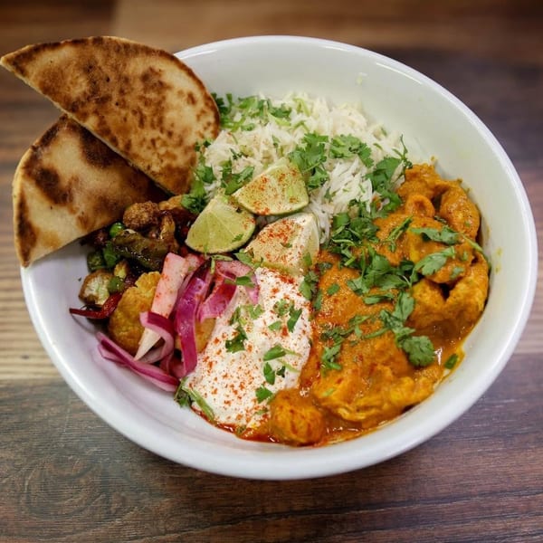 A vibrant Indian bowl with chicken curry, rice, vegetables, yogurt, lime, and naan bread.