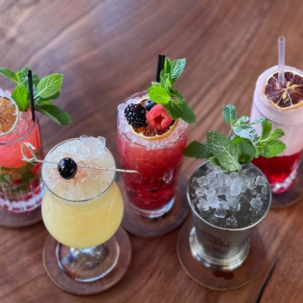 Five colorful cocktails with various garnishes like mint, berries, and dried citrus on a wooden table.