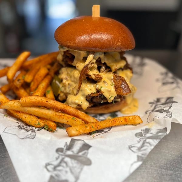 A towering burger with melted cheese and crispy bacon, served with seasoned fries on branded paper.