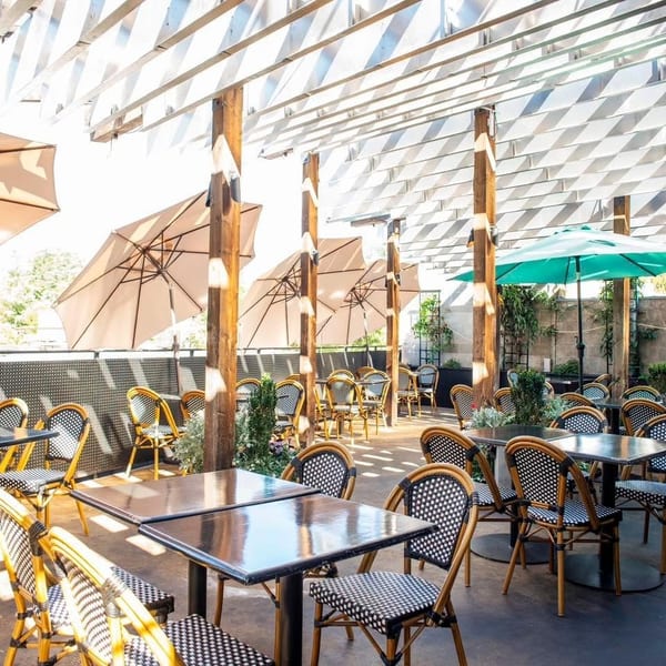 Sunny outdoor patio with black tables, wicker chairs, and umbrellas under a slatted pergola.