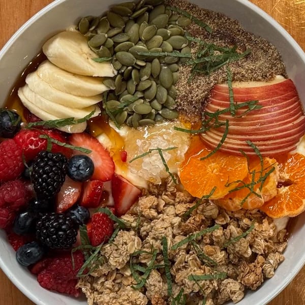 Overhead view of a colorful breakfast bowl with fruit, granola, seeds, and honey.
