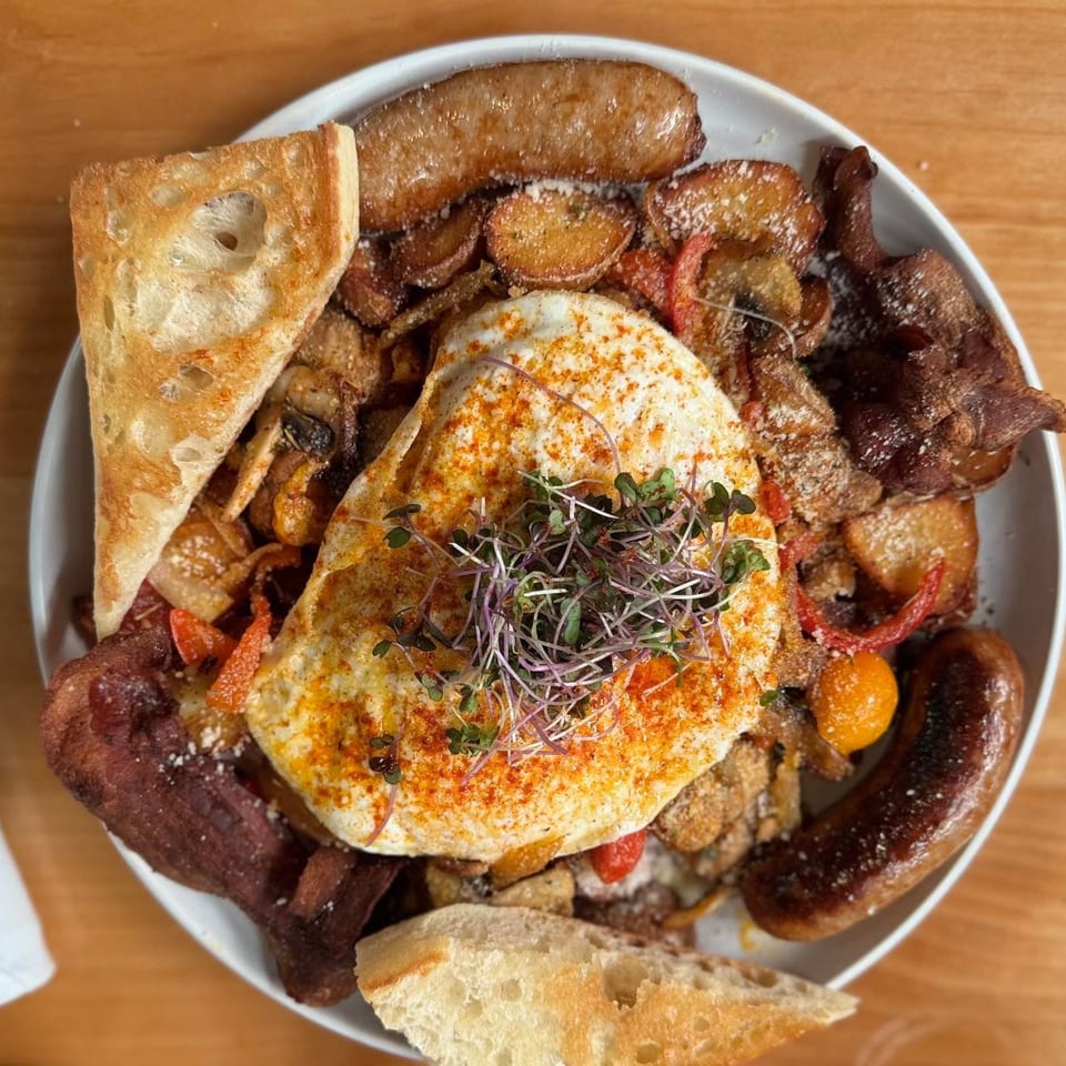 Hearty breakfast platter with fried eggs, sausage, bacon, potatoes, and toast on a wooden table.
