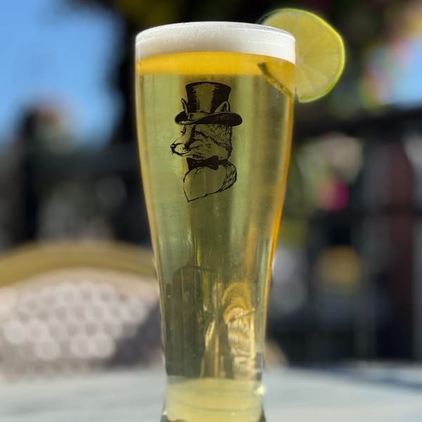A pilsner glass with a fox in a top hat logo, topped with foam and a lime.