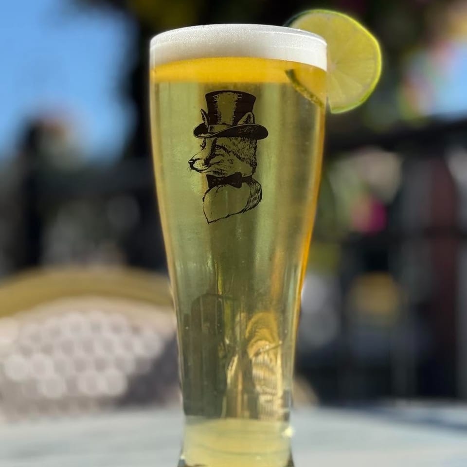 A pilsner glass with a fox in a top hat logo, topped with foam and a lime.