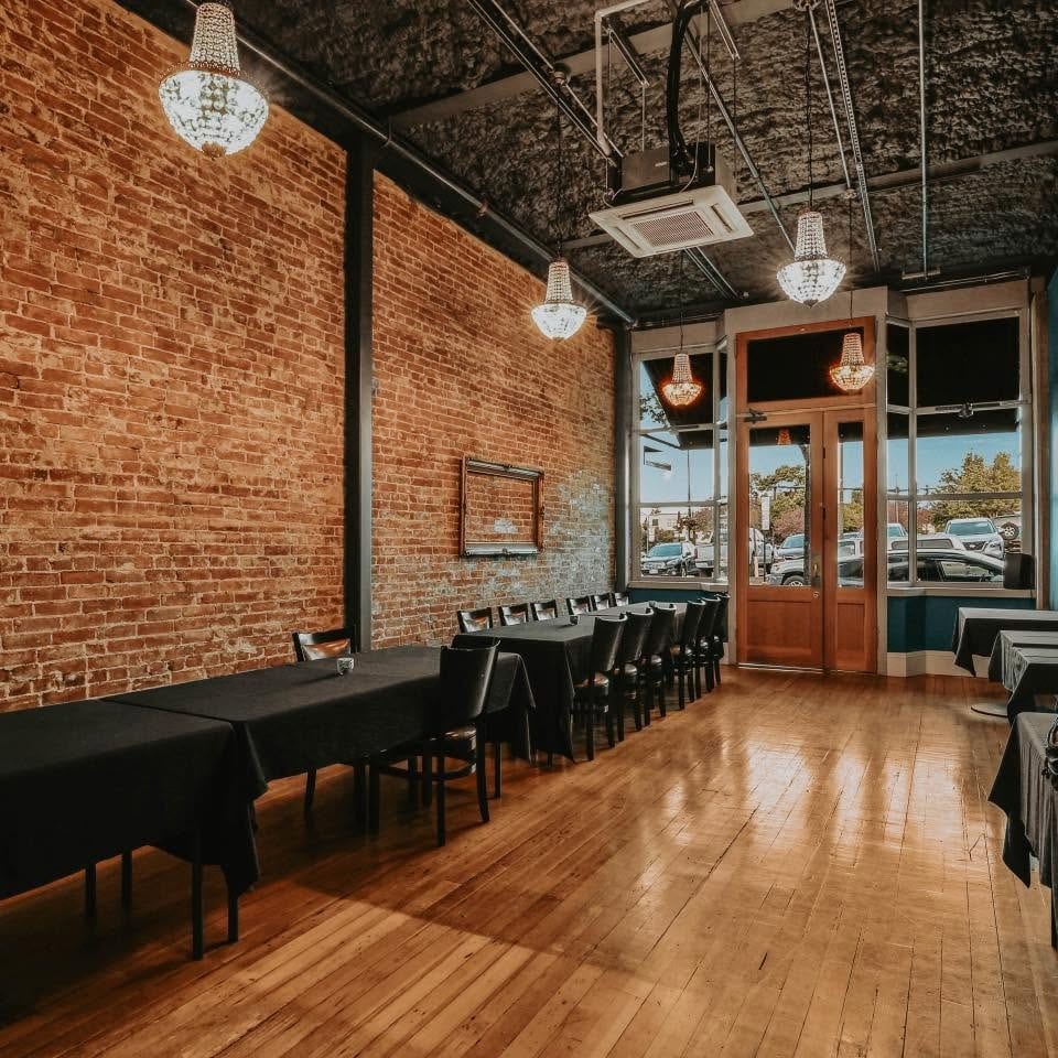 An empty event room with long tables, black tablecloths, brick walls, and chandeliers.