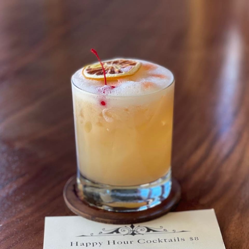 A foamy cocktail with a dried orange slice and cherry on a wooden coaster, next to a happy hour menu.